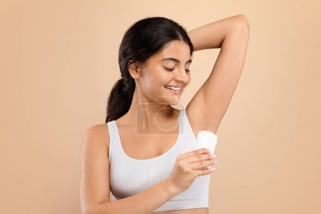 Photo for Personal Hygiene. Smiling Young Indian Woman Applying Deodorant Stick To Underarm Zone, Beautiful Eastern Female Using Antiperspirant On Armpit To Reduce Sweating, Posing Over Beige Studio Background - Royalty Free Image