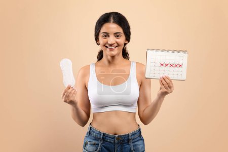 Photo for Smiling indian woman holding sanitary pad in one hand and a marked menstrual cycle calendar in the other, happy eastern female depicting womens health awareness, standing against beige background - Royalty Free Image