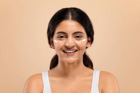 Portrait of cheerful young indian woman with white cream spots on her face smiling at camera, attractive eastern female enjoying skincare routine, standing against warm beige studio background