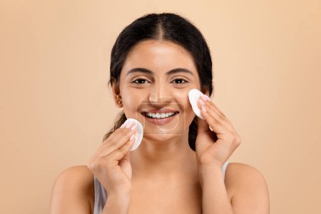 Portrait of happy young indian woman using cotton pads on her face with joyful expression, beautiful eastern female symbolizing refreshing skincare routine, standing against beige studio background