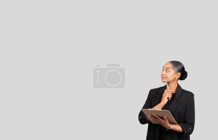 Photo for Thoughtful African American businesswoman holding a clipboard while looking upwards, contemplating new ideas against a plain grey backdrop, exemplifying professionalism and focus - Royalty Free Image
