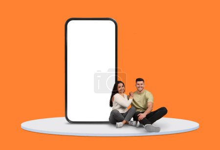 Lächelndes junges Paar im Schneidersitz auf dem Boden in der Nähe eines riesigen, leeren Smartphone-Bildschirms vor leuchtend orangefarbenem Hintergrund, perfekt für digitale Werbung