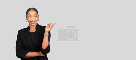 Foto de Confiada feliz millennial mujer de negocios afroamericana en traje profesional señalando a un lado con una sonrisa brillante y atractiva, sobre un fondo gris neutro, panorama - Imagen libre de derechos
