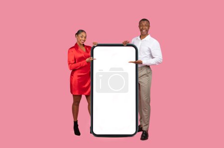 Téléchargez les photos : Homme noir enthousiaste en blanc et femme en robe rouge présentant une maquette géante de smartphone, parfait pour l'espace publicitaire, sur fond rose - en image libre de droit