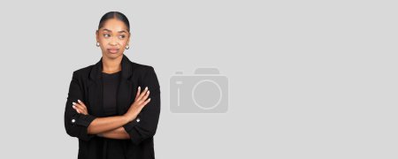 Téléchargez les photos : Femme d'affaires sceptique triste afro-américaine se tient avec les bras croisés, montrant un regard de curiosité et d'évaluation, placé sur un fond gris expansif, studio, panorama - en image libre de droit