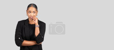 Foto de Mujer de negocios afroamericana pensativa mordiéndose las uñas, mirando hacia otro lado con una expresión preocupada, profunda en pensamiento o preocupación, sobre un amplio fondo gris, estudio, panorama - Imagen libre de derechos