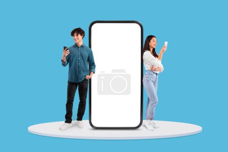 Foto de Joven hombre y mujer cada uno sosteniendo teléfono inteligente, de pie en lados opuestos de pantalla gigante en blanco vertical, contra el telón de fondo azul, maqueta - Imagen libre de derechos