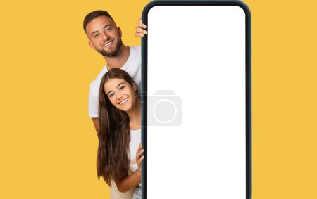 Téléchargez les photos : Un jeune couple européen ravi avec des sourires rayonnants pairs derrière un grand smartphone, symbolisant la connectivité et la communication moderne sur fond jaune, studio - en image libre de droit