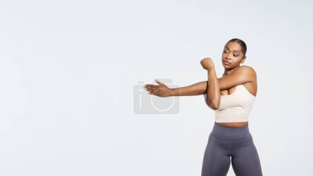 Téléchargez les photos : Femme de fitness afro-américaine faisant de l'exercice et étirant les bras avant l'entraînement sur fond de studio blanc. Ajustez femme en vêtements de sport échauffer les muscles. Panorama, espace libre pour le texte - en image libre de droit