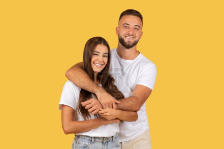 Foto de Una alegre pareja caucásica joven en camisetas blancas y vaqueros abrazándose y sonriendo ampliamente sobre un vibrante fondo amarillo, encarnando calidez y afecto. Amor, romántico - Imagen libre de derechos