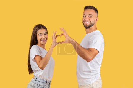 Téléchargez les photos : Heureux couple européen en t-shirts blancs en forme de cœur avec leurs mains, symbolisant l'amour et la connexion, avec des expressions joyeuses sur un fond de studio jaune. Amour, romantique - en image libre de droit