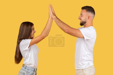 Photo for Energetic young couple in white t-shirts happily giving each other a high-five, celebrating teamwork and success against a bright yellow background. Spare time, fun together - Royalty Free Image