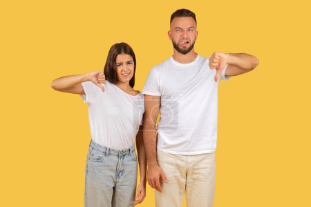 Photo for Disapproving young couple in white t-shirts giving thumbs down gestures with displeased expressions, indicating negative feedback or dissatisfaction against a yellow background - Royalty Free Image