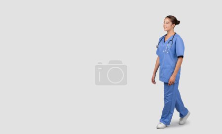 A confident and friendly Caucasian millennial healthcare worker in blue scrubs and stethoscope walking, exemplifying professionalism and approachability in medical attire