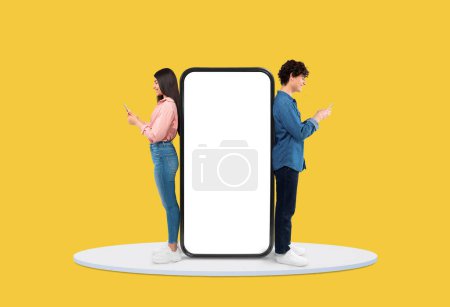 Teenage girl and boy engrossed in their smartphones standing back-to-back next to huge blank smartphone screen on yellow background