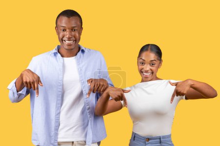 Téléchargez les photos : Avec des sourires brillants, l'homme et la femme noirs pointant vers le bas avec confiance, rayonnant de fierté et de positivité sur fond jaune ensoleillé - en image libre de droit