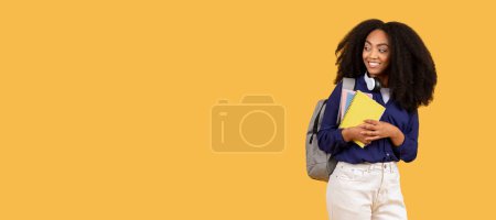Téléchargez les photos : Joyeux jeune femme noire aux cheveux bouclés et écouteurs, tenant des cahiers, regardant l'espace libre avec le sourire, debout sur un fond jaune chaud, panorama - en image libre de droit