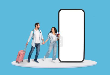 Téléchargez les photos : Joyeux jeune couple tenant la main et marchant avec leurs bagages de voyage à côté d'un écran publicitaire surdimensionné pour smartphone, isolé sur fond bleu - en image libre de droit