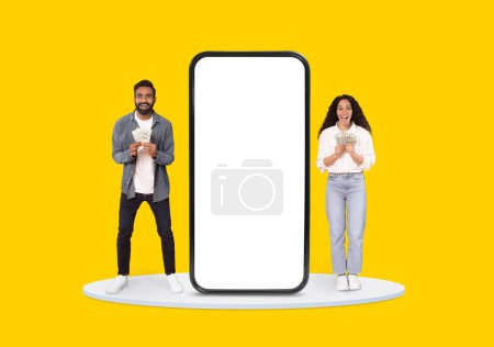 Téléchargez les photos : Homme et femme joyeux saisissant de l'argent à côté d'un écran de smartphone surdimensionné, suggérant le succès financier, sur fond jaune vif - en image libre de droit