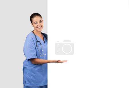 Foto de Invitando a la enfermera europea en uniformes azules que presentan un gesto de mano abierta junto a un espacio en blanco, ideal para publicidad sanitaria o visualización de información, aislado sobre fondo gris del estudio - Imagen libre de derechos