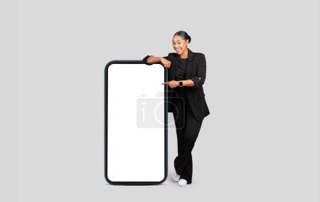 Foto de Joven y alegre mujer afroamericana en traje formal señala con el dedo el teléfono grande con la pantalla en blanco, aislado en el fondo gris del estudio, de cuerpo entero. Negocios, aplicación de trabajo, recomendación del sitio web - Imagen libre de derechos