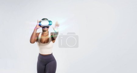 Téléchargez les photos : Femme afro-américaine active avec casque VR simulant des activités de gymnastique sur fond blanc, incarnant le mélange de fitness et de réalité augmentée, atteignant la main à la caméra. Espace vide, panorama - en image libre de droit