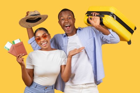 Téléchargez les photos : Heureux jeune couple noir tenant valise jaune, passeports et chapeau de paille, rayonnant de joie et d'anticipation pour leur voyage, sur fond jaune - en image libre de droit