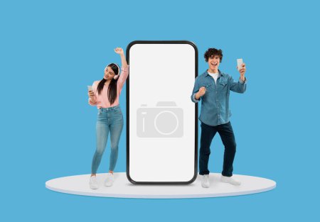 Photo for Happy teenage boy and girl dancing and enjoying music on their phones beside large vertical smartphone screen on light blue background - Royalty Free Image