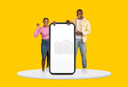 Photo for Cheerful black man and woman presenting large vertical blank smartphone screen, with ample copy space, isolated on vibrant yellow background - Royalty Free Image
