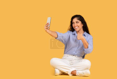 Photo for A cheerful young woman in casual clothes sits cross-legged on a yellow background, taking a selfie and giving a thumb up. Photo for social networks, approve gesture, recommendation - Royalty Free Image