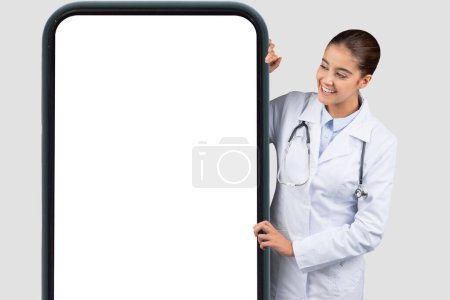 Photo for Cheerful Caucasian millennial doctor with a stethoscope appearing from the side of a large smartphone blank screen, perfect for telehealth or medical app promotions, blog - Royalty Free Image