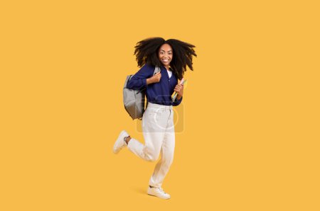 Foto de Alegre negro dama exudando entusiasmo, saltos en el aire con mochila y copybooks, conjunto sobre amarillo fondo, la captura de la esencia animada de la vida estudiantil - Imagen libre de derechos