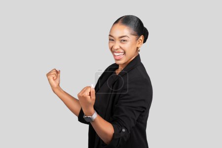 Téléchargez les photos : Exuberant heureuse femme d'affaires afro-américaine millénaire avec un sourire rayonnant, vêtue d'un élégant costume noir, célébrant le succès avec une pompe de poing jubilant sur un fond neutre, studio - en image libre de droit