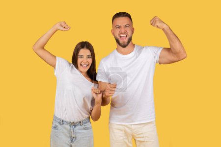 Photo for Excited young couple in white t-shirts showing off their muscles and celebrating success with a cheerful attitude against a yellow background. Winner gesture, team victory together - Royalty Free Image