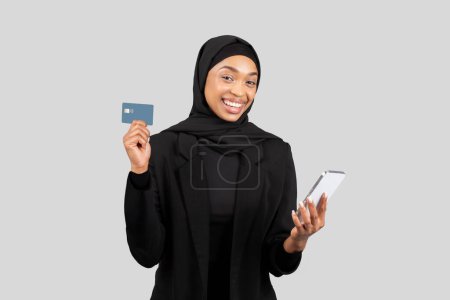 Foto de Alegre mujer de negocios afroamericana en hijab sosteniendo una tarjeta de crédito y un teléfono inteligente, retratando la facilidad de las transacciones digitales y la independencia financiera, aislado en el fondo gris del estudio - Imagen libre de derechos