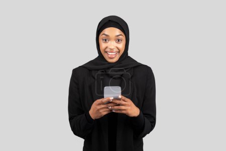 Photo for Smiling surprised millennial black lady in hijab chatting on smartphone in social networks, isolated on gray studio background. Business, work app, communication online, read message - Royalty Free Image
