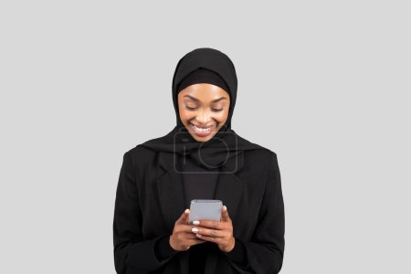 Foto de Mujer de negocios afroamericana sonriente en hijab interactuando con un teléfono inteligente, que representa profesionalismo moderno y conectividad en el lugar de trabajo, aislado en el fondo del estudio gris - Imagen libre de derechos