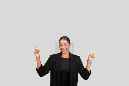 Foto de Alegre mujer de negocios afroamericana apuntando hacia arriba con ambas manos, mostrando un producto o idea con una sonrisa brillante y atractiva contra un fondo gris, estudio. Anuncio de trabajo, venta - Imagen libre de derechos