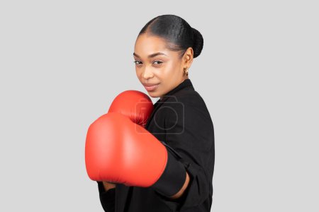 Téléchargez les photos : Femme d'affaires afro-américaine confiante en costume noir avec des gants de boxe rouges, représentant l'autonomisation, la force et la concurrence dans le monde des entreprises, isolée sur fond de studio gris - en image libre de droit