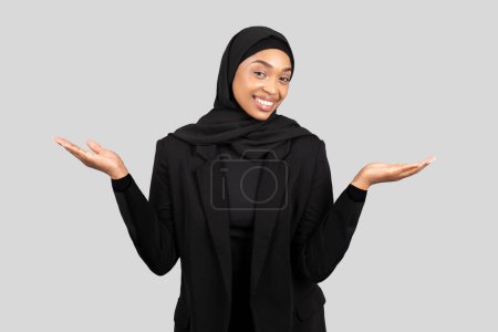 Foto de Glad millennial empresaria negra en hijab mantener en ambas manos espacio libre, lo que sugiere ideas innovadoras y objetivos, aislado en fondo de estudio gris. Elección, anuncio y oferta - Imagen libre de derechos