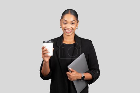 Téléchargez les photos : Femme d'affaires afro-américaine souriante tenant une tasse de café et un ordinateur portable, prête pour une journée productive, combinant la technologie et un boost de caféine dans un costume noir sur fond gris, studio - en image libre de droit