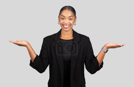 Photo for Balanced glad African American businesswoman with open palms facing up, presenting two options or weighing choices with a friendly smile in a professional setting on grey background, studio - Royalty Free Image