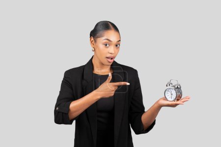 Téléchargez les photos : Une femme d'affaires afro-américaine surprise en costume noir pointe un réveil qu'elle tient, symbolisant la gestion du temps, les délais et la ponctualité dans un environnement d'entreprise - en image libre de droit