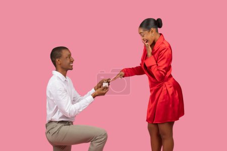 Foto de El hombre negro se arrodilla con la caja abierta del anillo que propone a la mujer extática en vestido rojo vibrante, su mano sobre su boca en alegría, sobre fondo rosa - Imagen libre de derechos