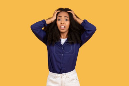 Photo for Stressed young black woman with curly hair looking anxious, her hands on her head, standing against plain yellow backdrop expressing concern - Royalty Free Image