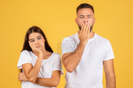 Photo for Young woman with a bored expression resting her cheek on her hand and man covering his mouth with hand in a shocked gesture, both wearing white t-shirts against a yellow backdrop - Royalty Free Image