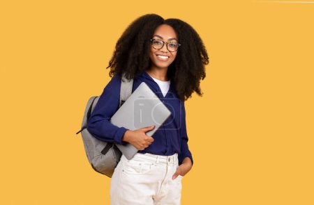 Téléchargez les photos : Étudiante noire excitée portant un sac à dos, tenant joyeusement un ordinateur portable et souriant directement à la caméra, sur fond jaune vif, incarnant l'enthousiasme dans la technologie et l'apprentissage - en image libre de droit