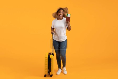 Téléchargez les photos : Heureuse touriste noire tenant billet d'avion et passeport debout avec sa valise sur fond de studio jaune, pleine longueur prise de vue avec espace vide pour l'offre de l'agence de voyage - en image libre de droit