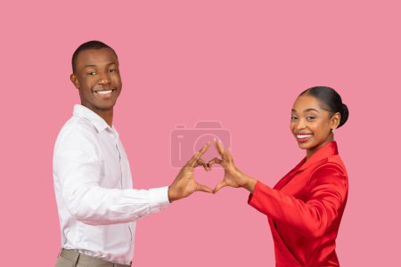 Téléchargez les photos : Heureux couple noir en tenue élégante créant forme de coeur avec leurs mains ensemble sur fond rose, symbolisant l'amour et souriant à la caméra - en image libre de droit
