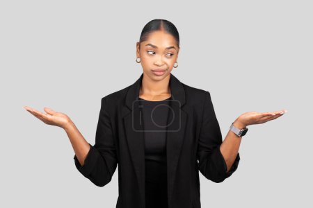 Photo for A skeptical millennial African American businesswoman in a black suit standing with open hands, weighing options with a doubtful expression on a gray backdrop, studio - Royalty Free Image